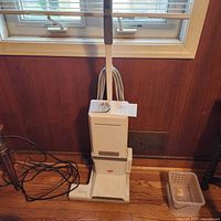 Front view of the upright vacuum cleaner showing the base, handle, cord, and overall shape in white with slightly worn condition.