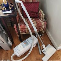 Full view of white Electrolux Lux Classic canister vacuum with hose, metal wand, and floor head attachment placed on wooden floor near chair.
