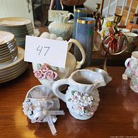 Porcelain vases with 3D flower blooms, some smaller decorative ceramics, tall blueish gray glass vase, and a metal horn with ribbon bow on wooden table.