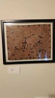Full view of the framed tribal art showing abstract figures and animals on brown vinyl mat.