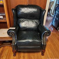 Full front view of dark green leather wingback recliner chair with brass nailhead trim, sitting on wooden floor next to wooden furniture.