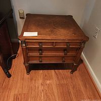 Front view of walnut MCM end table showing 3 drawers, wood grain, round drawer pulls, and brass details on legs and corners.