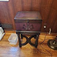 Closed view of dark wood pedestal chest with decorative crest on front panel, showing turned leg base on hardwood floor.