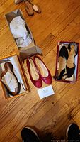 Three pairs of size 10 women's shoes on the floor: beige leather sandals in a box, red ballet flats, and black strap flats in a box.