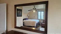 Photo of large rectangular wall mirror with dark wood frame reflecting bedroom.
