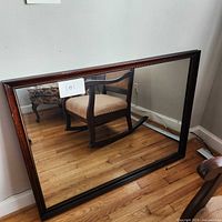 Full view of the wood framed mirror reflecting a chair and wooden floor, showing size and overall condition.