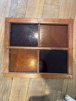 Top view of square wooden window frame with four colored glass panels in dark purple, brown, and amber shades arranged in a 2x2 grid, placed on a wooden floor.