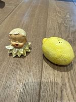 Photo of two salt and pepper shakers, a cherub head figure and a ceramic lemon on a wooden surface.