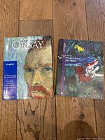 Photo of two art books laid flat on a wooden floor: the left book is 'Your Visit to Orsay' featuring Van Gogh's portrait on the cover; the right book is 'Album Message Biblique Musée Chagall' with a colorful artistic cover.