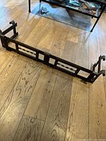 Brass antique fireplace grate on wooden floor showing full top-down perspective with curled end details and front geometric panels