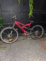 Side view of a red Supercycle mountain bike with visible branding and suspension fork, parked outdoors on pavement in front of a dark metal gate, surrounded by some greenery.