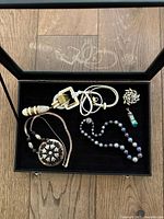 Black interior of jewelry case containing three necklaces, one brooch, and one pendant.