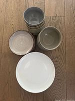 Set of dishes and bowls placed on wooden floor showing white plates, light earth tone bowls stacked, matching side plates with a circular pattern, and one large white serving plate.
