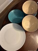 7 stacked blue ceramic bowls next to one white dinner plate and one blue ceramic plate on wooden surface