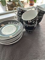 Six black espresso cups with white and red coffee text stacked and six matching black saucers with white 'espresso' text and coffee bean graphics on a grey surface.