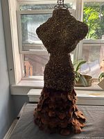 Back and side view of wire framed mannequin torso covered in textured material with layered feather-like skirt, shown near window for natural light