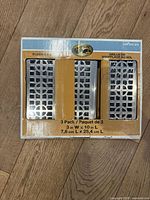 Photo of the set of three floor vent grates in original packaging, showing the metal grates through the box windows with decorative cutout patterns, size 3 by 10 inches each.