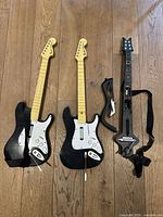 Three Guitar Hero controllers laid on wooden floor, two black and white and one black with colored fret buttons and strap.
