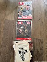 Photo showing two LIFE magazine issues on wooden floor with other loose pages and magazines beneath, some visibly worn with torn covers and edges.