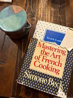 Photo showing Mastering the Art of French Cooking Volume Two book and a pottery butter holder on a wooden surface. Dust jacket of book has visible tear at upper right corner.