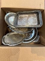 Top view of assorted silver plate and pewter trays in cardboard box showing tarnishing and wear.