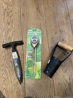 Photo showing three gardening products: a moisture, light and pH meter in its green packaging, a metal garden hand tool with a wooden handle and a pointed blade, and a metal hand cultivator tool with wooden handle.