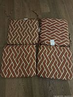 Four outdoor seat cushions arranged in a 2x2 grid on a wooden floor showing a brown and white geometric pattern, fabric ties attached for securing.