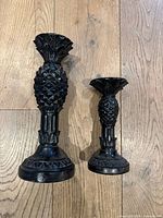 Photo showing two black wooden pineapple-shaped candlesticks of different sizes on a wood floor.