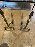Pair of vintage brass andirons standing on wood floor, showing full front view and base design
