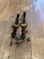 Pair of vintage brass andirons standing upright on a wood floor, tops showing wooden finials and brass bases with patina.