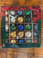Photo of a set of 25 vintage glass Christmas ornaments in their original sectioned box. The ornaments show a variety of metallic colors and finishes including green, gold, blue, silver, and red.