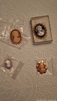 Four cameo brooches arranged on a white lace fabric surface, showing various sizes and styles.