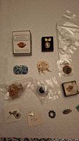 Lot of assorted brooches and pins laid out on a beige textured surface, showing various items in small plastic bags and boxes.