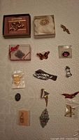 Photo showing 15 assorted brooches in various designs and materials including boxed and loose pieces arranged on a white textured background.