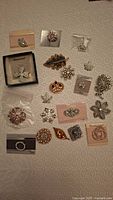 Top view of 20 assorted brooches arranged on a white fabric background showing variety of designs and styles.