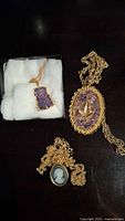 Three necklaces laid out: two amethyst pendant necklaces and one black and white cameo pendant necklace, all with gold-tone chains, one amethyst necklace in jewelry box