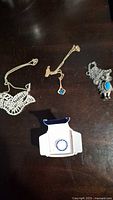 Photo of four necklaces on a dark surface showing two blue stone pendants, one beaded wing pendant, and one packaged necklace with blue accents