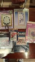 Photo showing the six sewing kits laying flat, various packaging with floral and bird cross-stitch designs, including Bucilla and Janlynn kits.