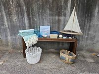 Full setup of varied beach-themed decor arranged on and around a wooden bench including glasses, bottles, artwork, bowls, seashell candy dish, throw blanket, baskets, and wooden sailboat model.