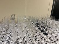 Wide view showing an extensive mixed set of vintage glassware on a crocheted tablecloth, including multiple types of glasses with varied etched and cut patterns, and different stem colors such as clear, black, and gold.