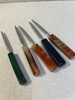 Photo shows five metal letter openers with polished agate stone handles in a variety of natural colors, laid flat on a beige surface.