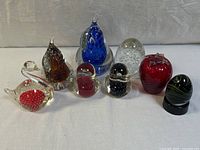 Eight decorative art glass paperweights on neutral background showing various shapes and colors including fruit shapes, swan, and abstract forms.