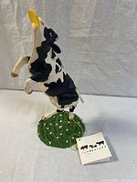 Side view of black and white cow figurine jumping with yellow frisbee, showing grassy base and attached CowParade hang tag.