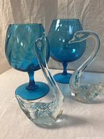 Front angle view of two clear glass swans with suspended blue flecks plus two blue glass goblets, one with swirl and one with smooth texture.