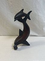 Front view of the hand-carved wooden sculpture showing two dolphins with smooth dark finish and natural wood grain, larger dolphin holds a black spherical ball in its mouth.