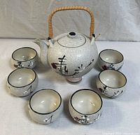 Round ceramic teapot with woven bamboo handle surrounded by six matching cups with white crackle glaze and hand-painted decorative motifs.