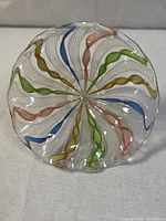 Top view of the Murano-style art glass dish showing clear glass with swirling green, blue, pink, and gold ribbons forming a symmetrical pattern radiating from the center.