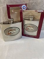 Two stainless steel hip flasks with Denver Broncos logo shown, one new in box and one out of box