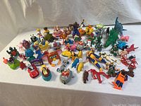 Full view of the entire toy collection laid on white fabric showcasing the varied colors, characters, and types of toys including vehicles and figurines.