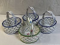 Four hand-painted ceramic lattice baskets arranged on a white fabric surface, showing three blue trimmed and one green trimmed baskets.